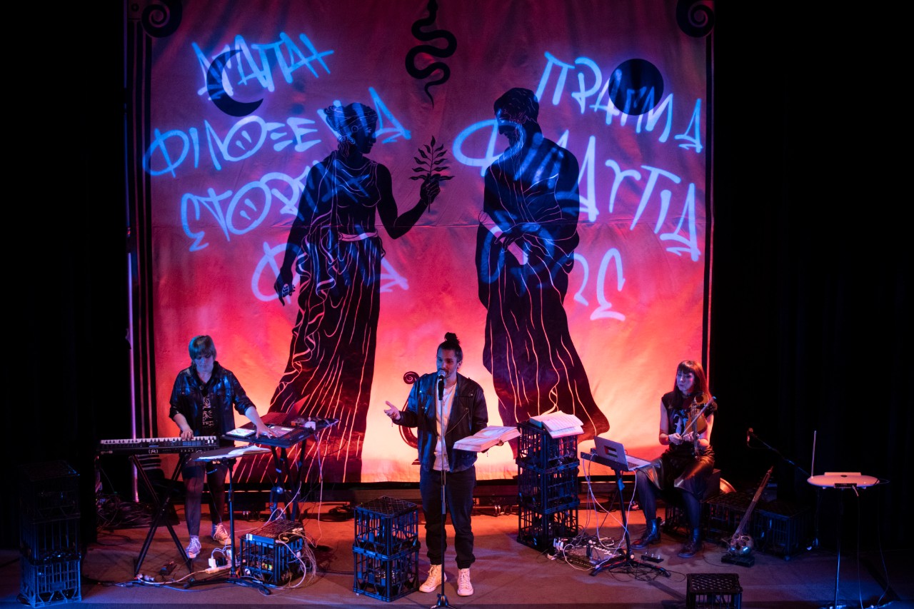 a stage setting in a theatre, one man is at the microphone speaking with two musicians standing behind him.