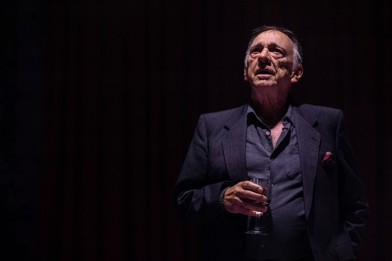 a actor is on stage, he is an older man holding a glass of beer