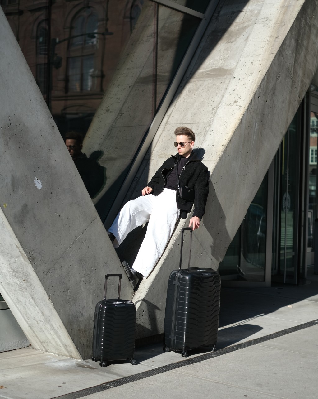 Pasha leaning against a diagonal pillar whilst lightly holding a suitcase