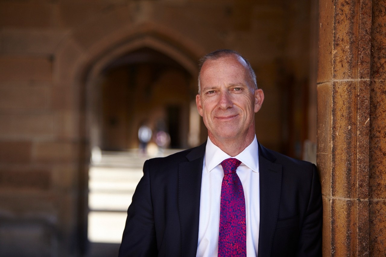 Dr Michael Spence leanign against a sandstone wall