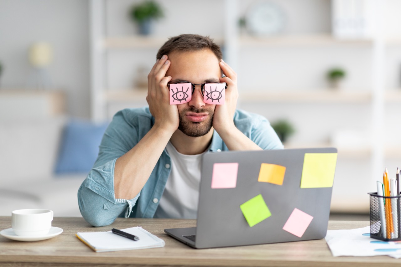 A man sleeping at his work desk with sticky notes covering his eyes with drawings of open eyeballs on them