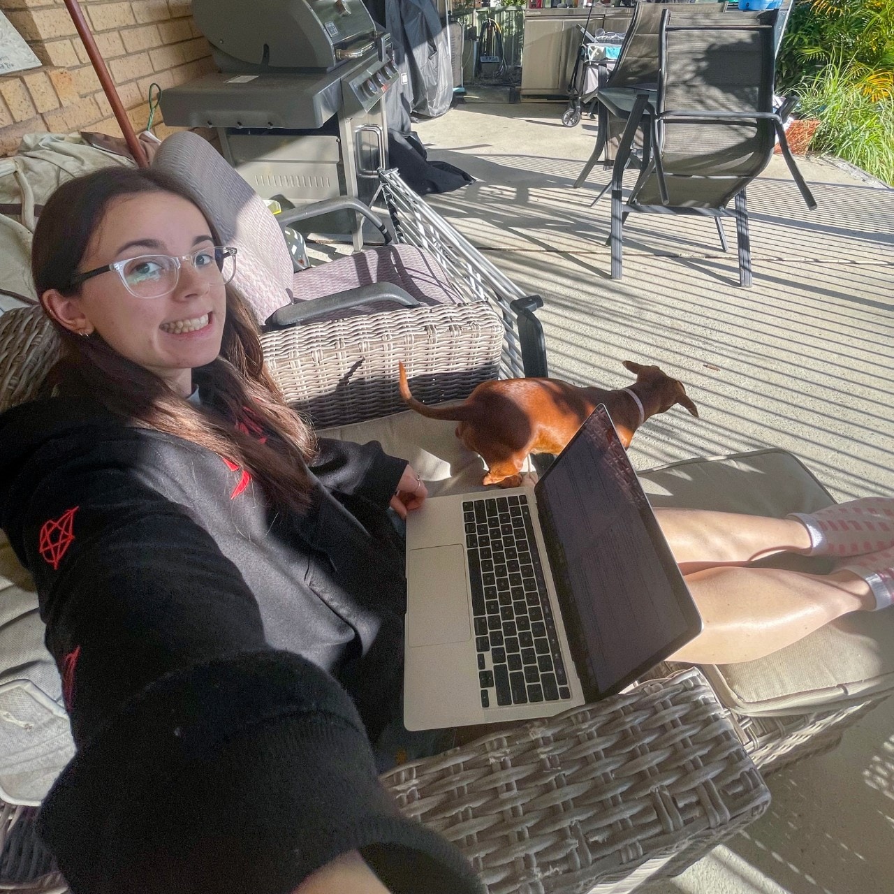 Cameryn Smider sits on a chair on an a deck outside her home with her laptop, smiling at the camera with a dog nearby.