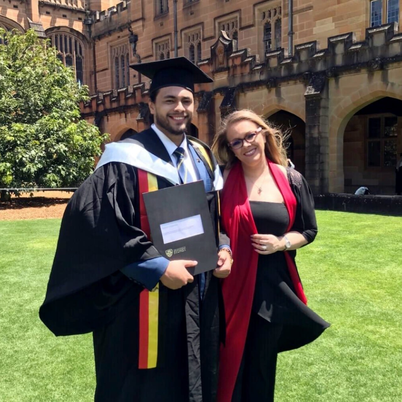 Patrick Lucarnus in his graduation holding his degree certificate, standing with a woman, on the grass in the University of Sydney's Main Quadrangle.