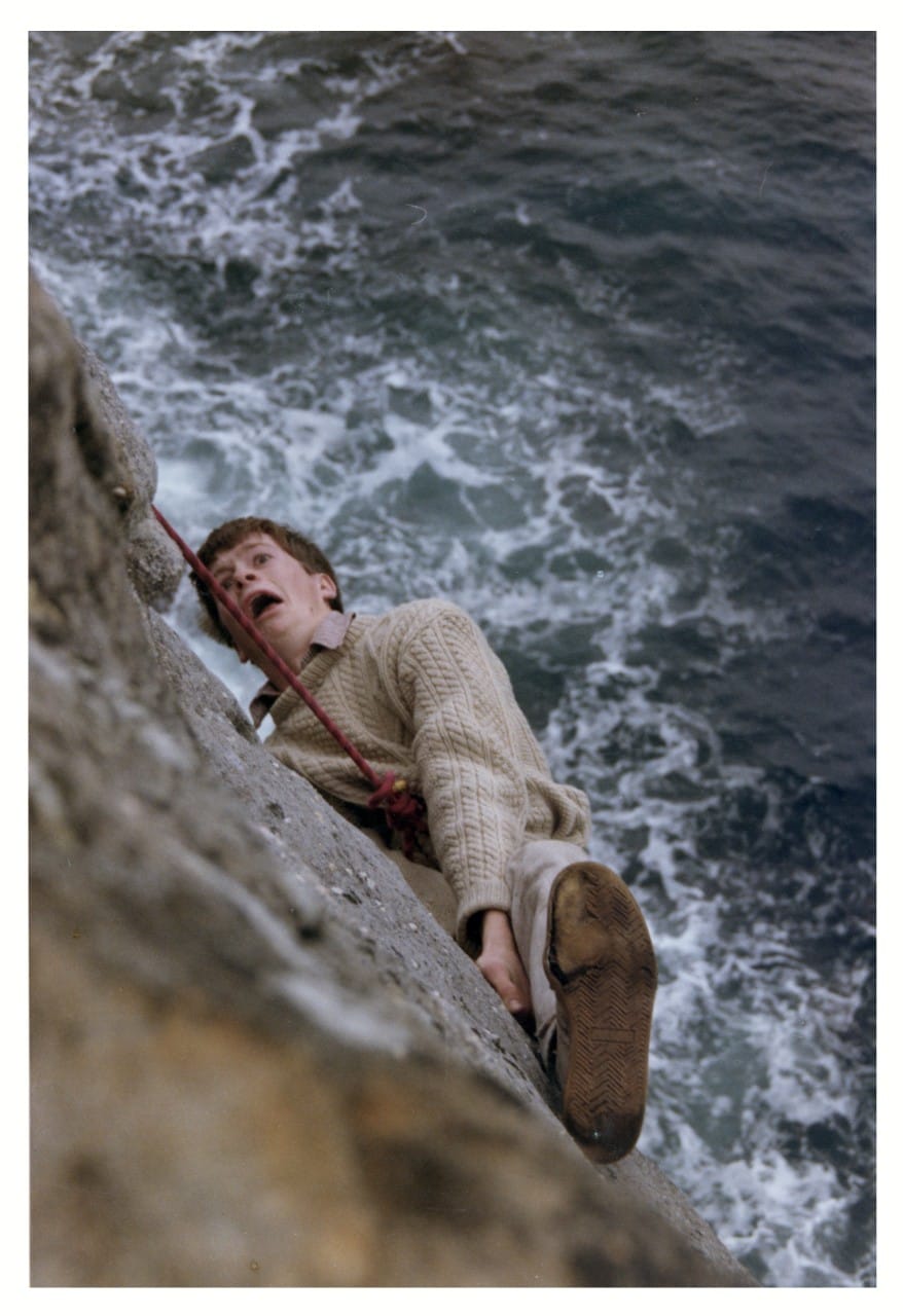 A student rock climbing on a cliff above the ocean appears to be in a precarious yet comic situation.