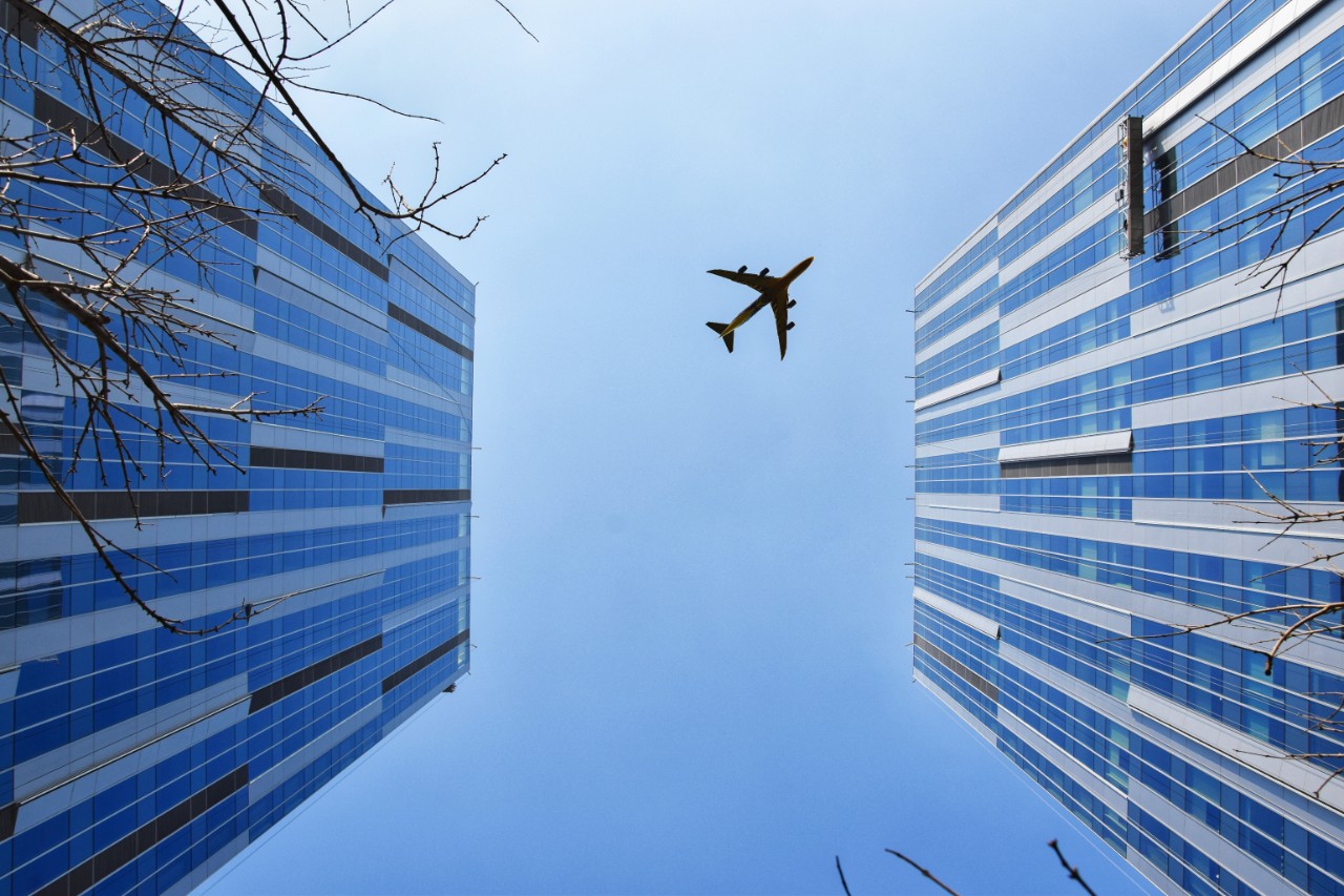 Image of plane flying through blue sky with two buildings on either side