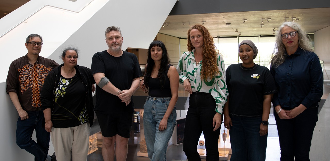Seven artists standing in a line in front of a railing, smiling.
