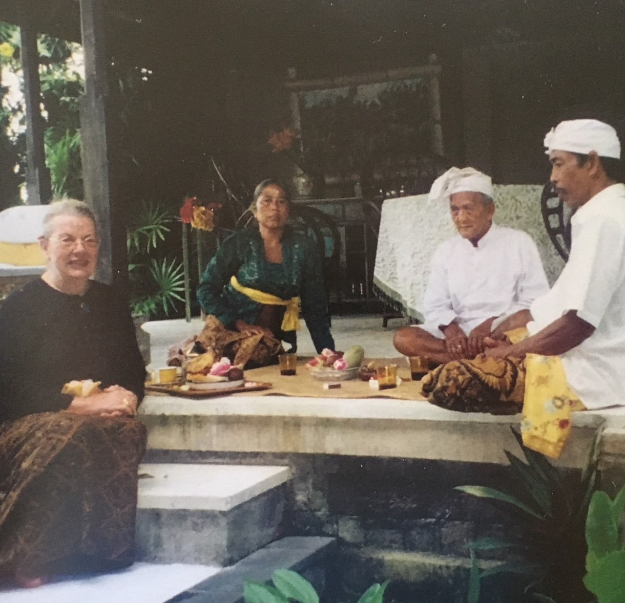 Bespectacled woman in black long-sleeved top and long patik-printed skirt sitting on a step with a couple plates of fruits and small cups of tea on a rattan mat, next to three local Balinese including a woman in traditional Balinese outfit of long-sleeved dark green top and batik skirts and two men with white head-wraps and long-sleeved tops and batik skirt.