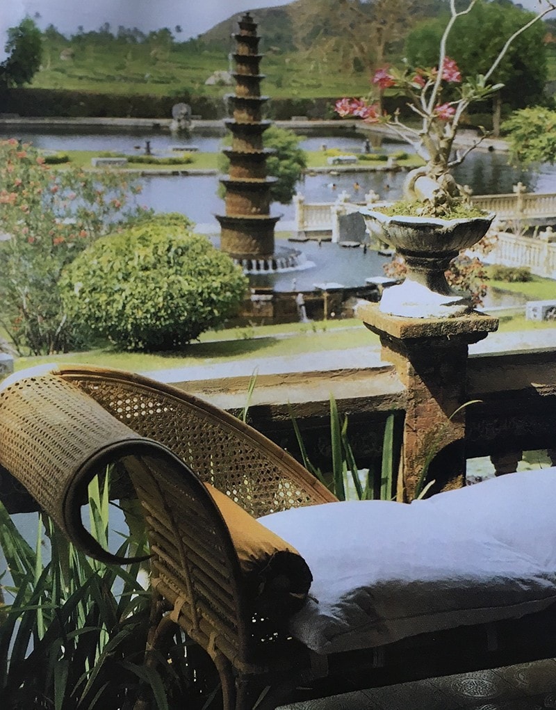 Lounge day-bed with rattan-like curved head-rest in the foreground, with lush green water garden fountain with small padoga-shaped waterfall structure in the centre