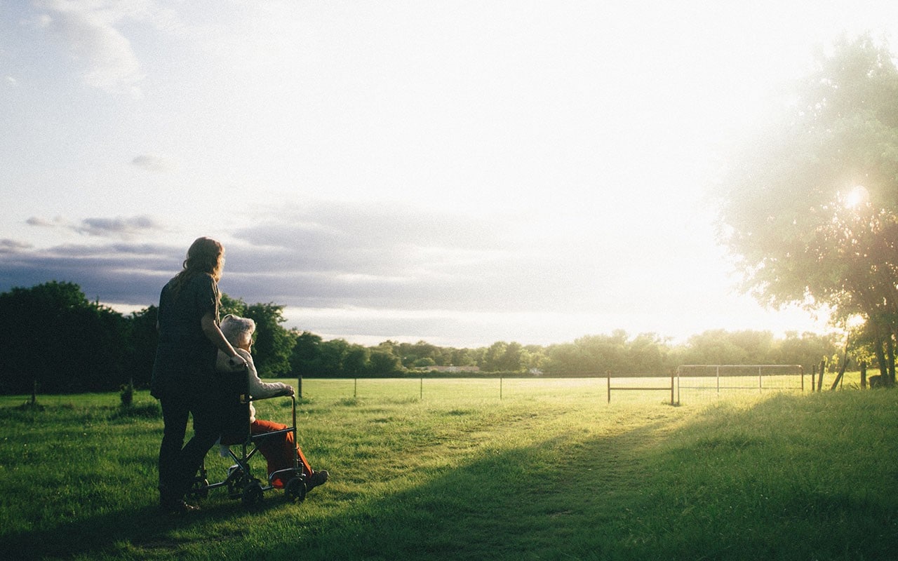 image showing standing woman with elderly person in wheelchair in grassy field looking toward setting sun