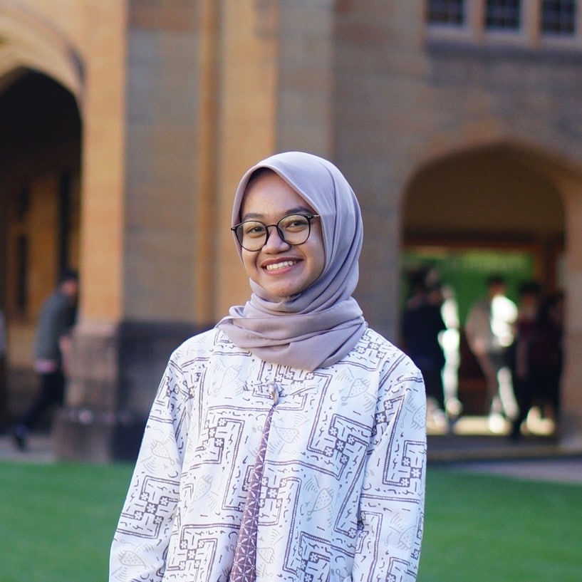 Alisha Nurul Fatiha is wearing a lilac hijab and a long sleeve, white shirt with a lilac square pattern. She has black glasses and is standing in front of a major building at the University of Sydney. She is smiling.