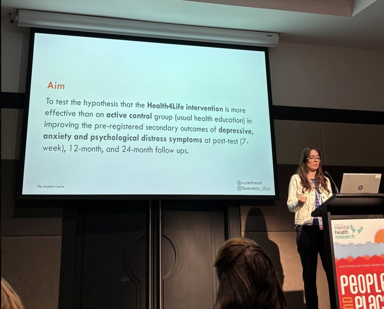 Scarlett presenting at SMHR. She is standing behind a podium and has a slide behind her, discussing the aims of the Health4Life program. 
