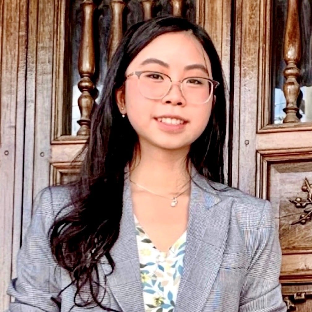 Stella Le is wearing a white patterned shirt with a grey blazer over the top. She is wearing thin, black glasses and has long, dark hair. She is smiling. 