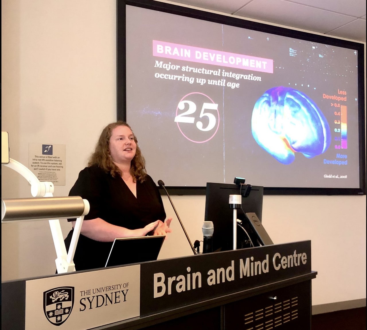 Steph presenting at a conference. She is standing behind a podium, and there is a slide in the background discussing the Illicit Project, a brain relationship program Steph leads.