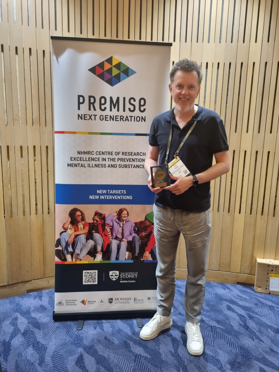 A man wearing a brown jumper and jeans holds a metal next to a sign saying 'PREMISE'