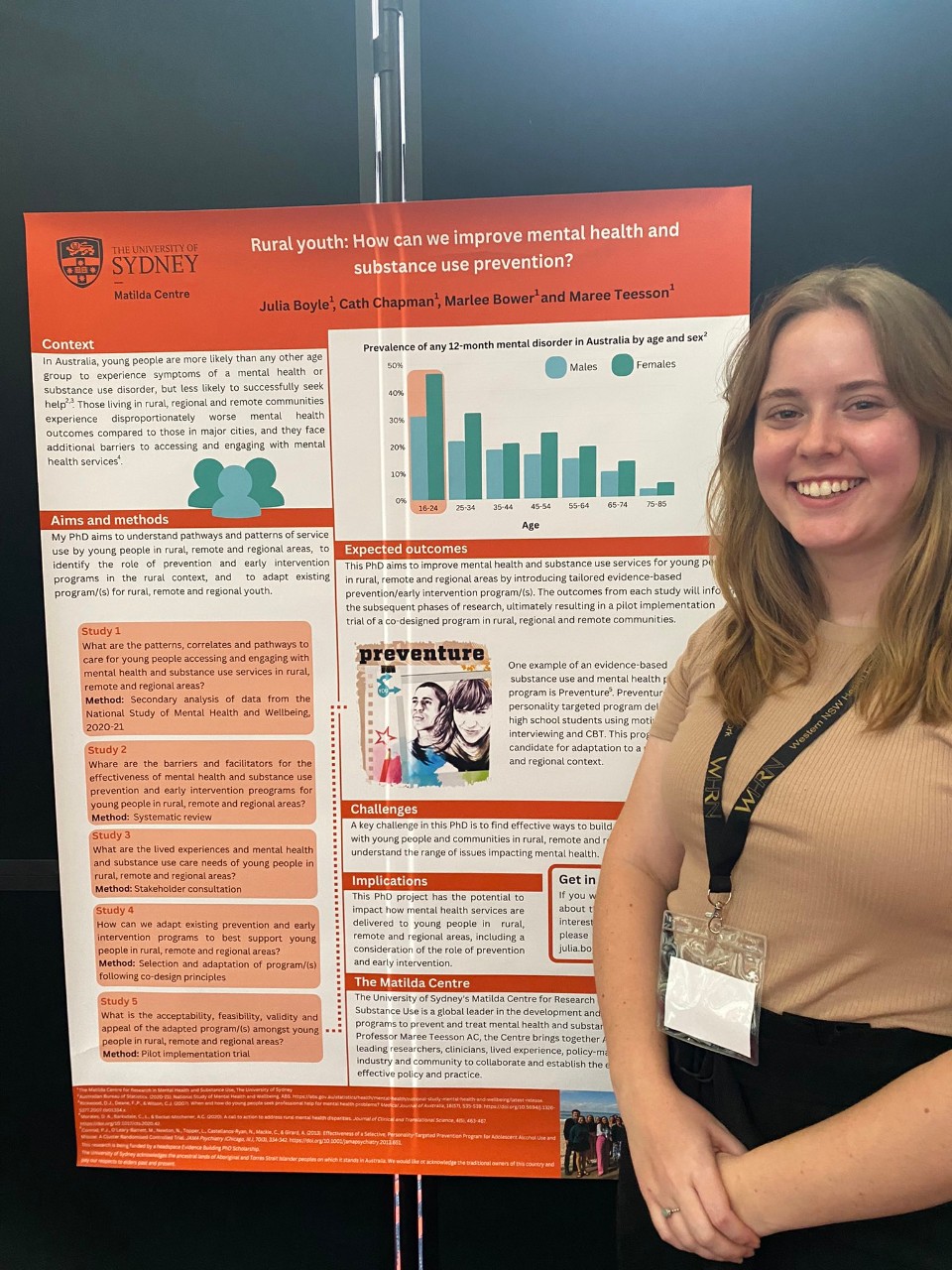 Julia is standing in front of a poster. She has long brown hair that she is wearing out and is wearing a white shirt. The poster behind her discusses her research into regional communities. 