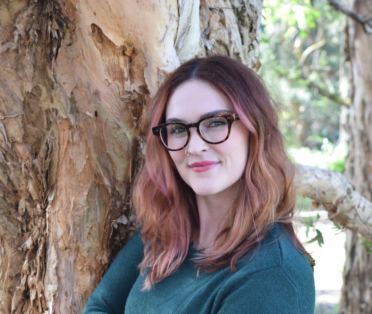 Siobhan is leaning against a tree and is wearing a dark green cardigan. She has dark, thick glasses and has shoulder length red hair. She is smiling