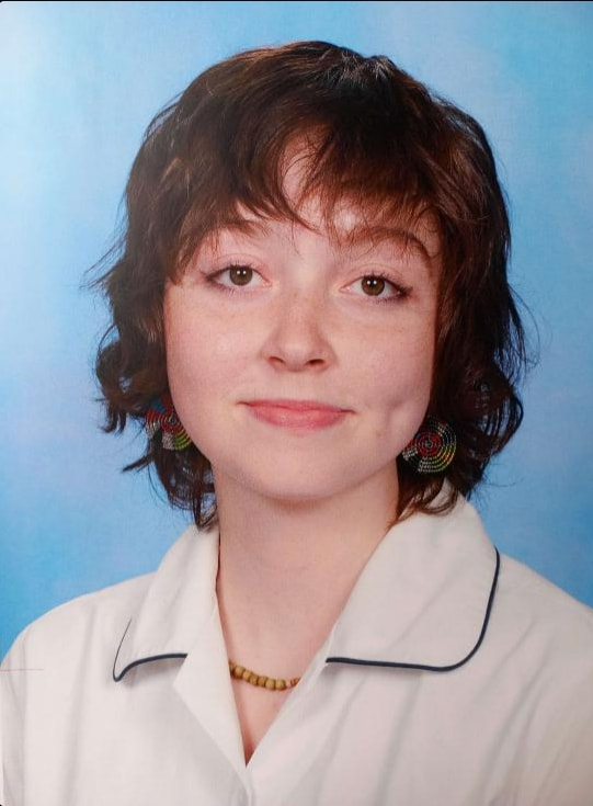 A headshot of Kyra. Kyra has short, dark hair and is wearing a white button up shirt with black lining. 