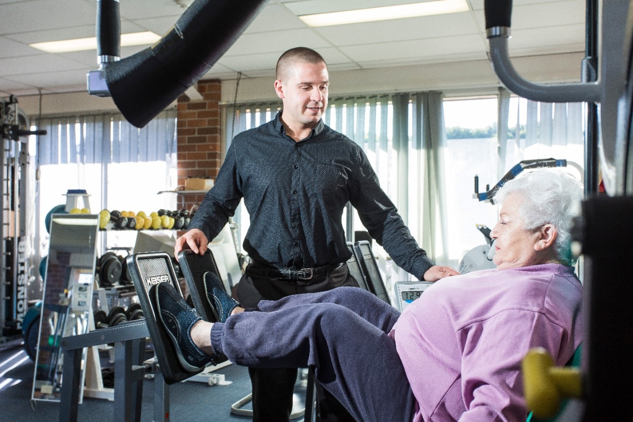 HDR student Michael Inskip instructing elderly woman doing leg presses on a weights machine in a gym