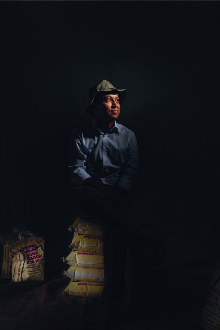 Shaymal Chowdury wearing a cap and in a darkened space with his face and other touch points gently illuminated. He is sitting on sacks of rice.