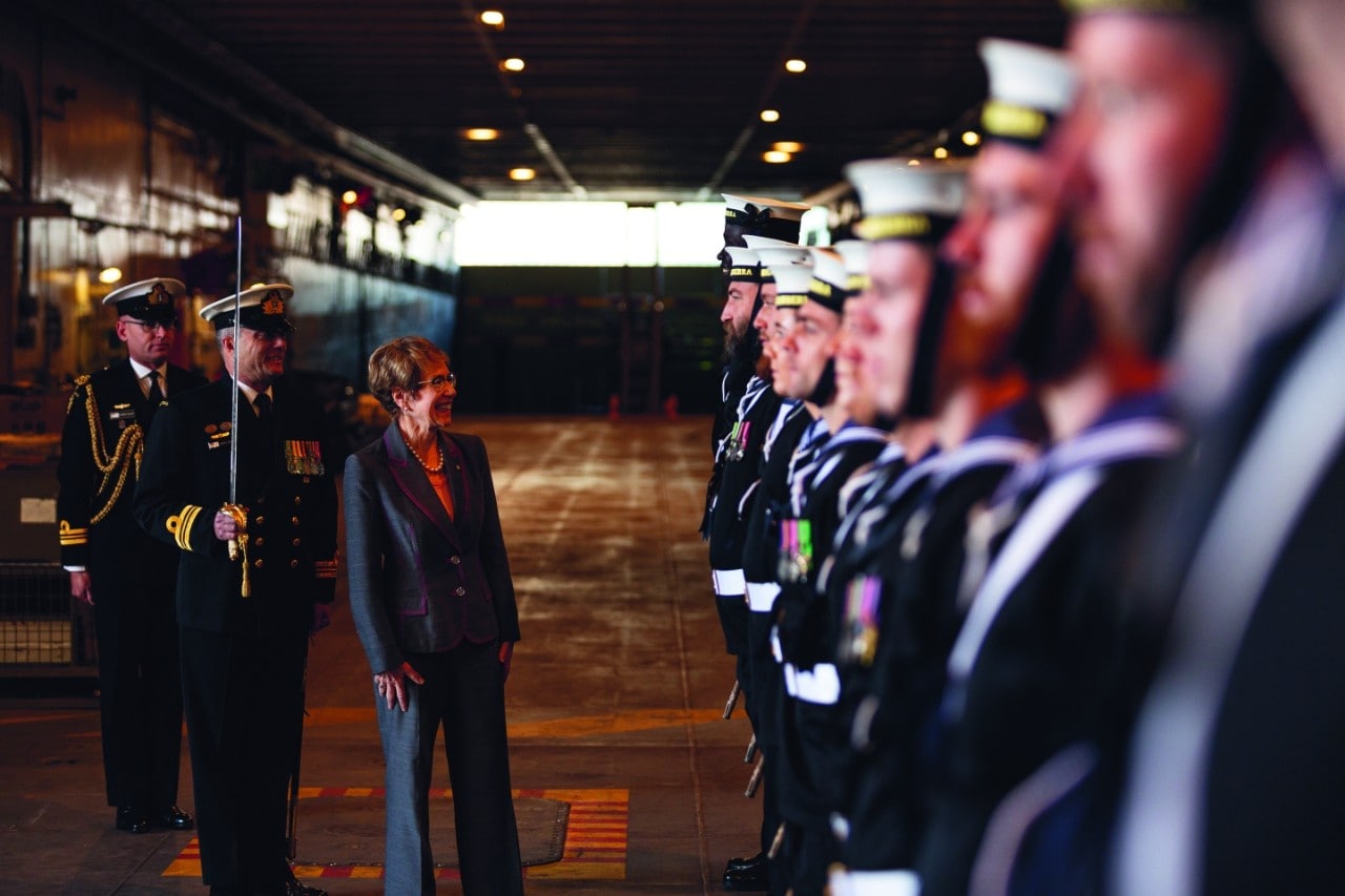 On the below deck of a navy ship and in low light, Beazley is inspecting a squad of sailors. She is on the left of frame with two officers dressed in ceremonial uniforms, one holding a sword. She is looking at a row of sailors on the right, with the closest to the camera blurring into the frame.