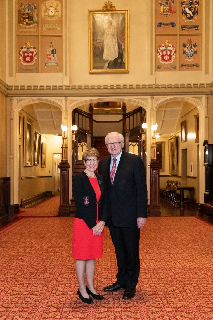 Margaret Beazley and Dennis Wilson posed standing together in the formal entrance foyer of NSW Government House. Behind them is a grand staircase and there is a sense of height and space. The carpet is red, and the walls seem golden being adorned with two crest banners and an old portrait in the centre.
