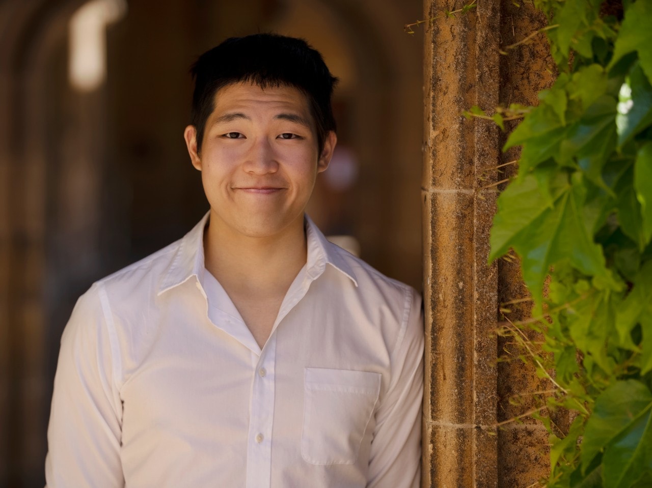 Joshua Wan wears a white shirt and stands beside a sandstone wall covered in ivy.