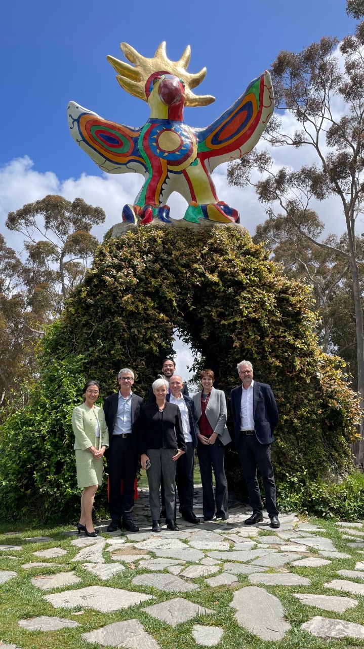 University of Sydney delegation stand with staff from UC San Diego in front of a large arch-like sculpture, covered in vines with a colourful bird-like head on top