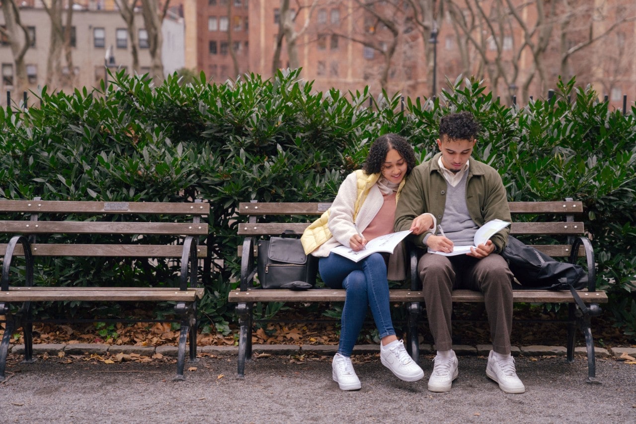 Two students sit on a park bench in the city comparing notebooks.