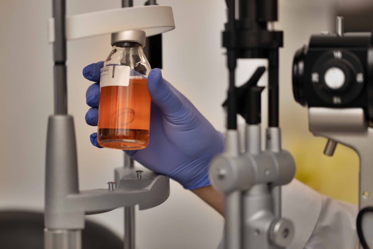 A photo of a gloved hand holding a specimen jar with a pale orange liquid, suspended within is an artificial cornea