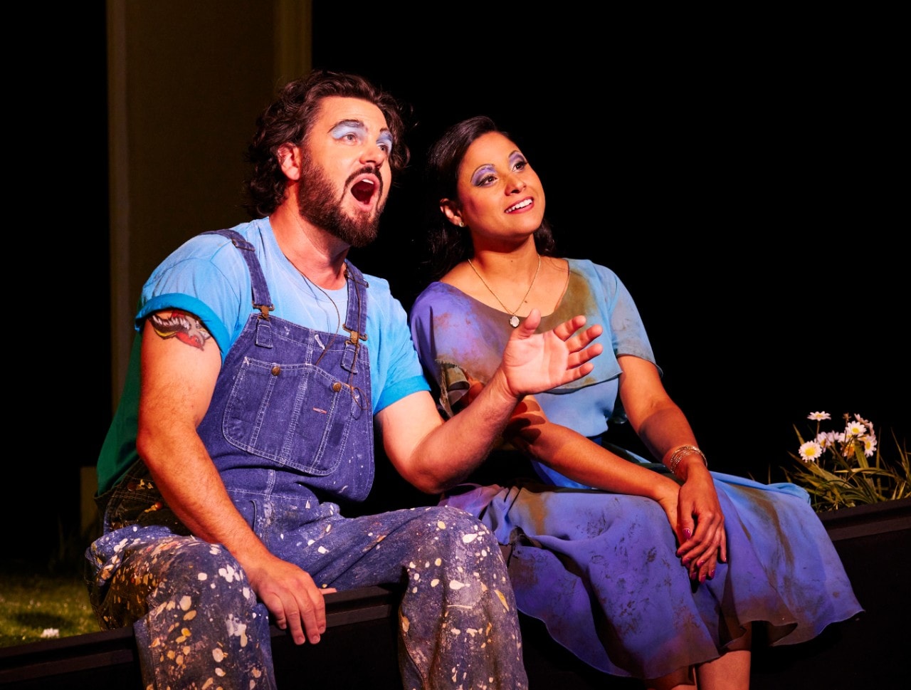 A man and a woman both wearing blue overalls sing to each other