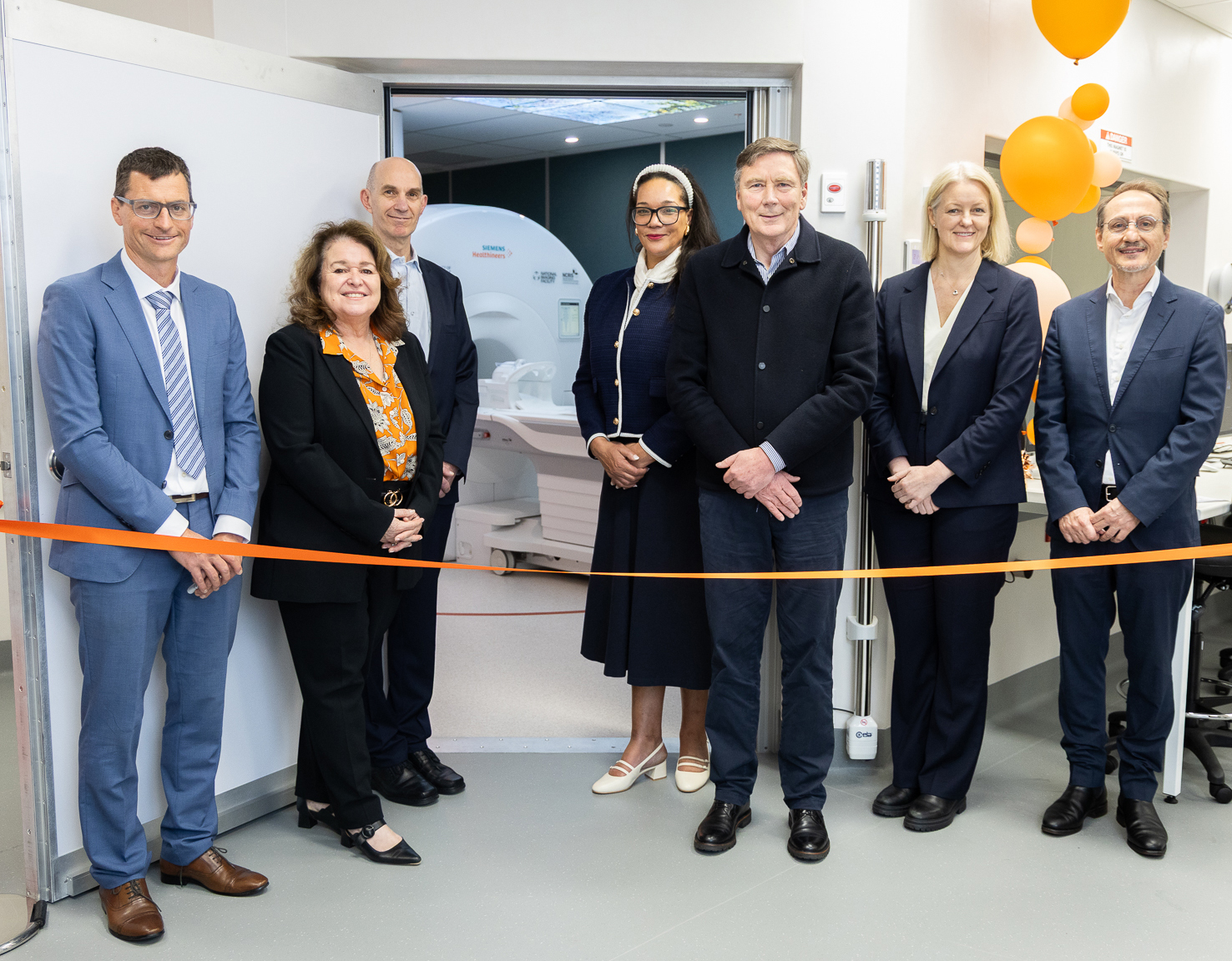 Photo of the 3T MRI facility ribbon cutting. Executives from the University of Sydney and Siemens Healthineers stand behind an orange ribbon with the new MRI machine in. the background.