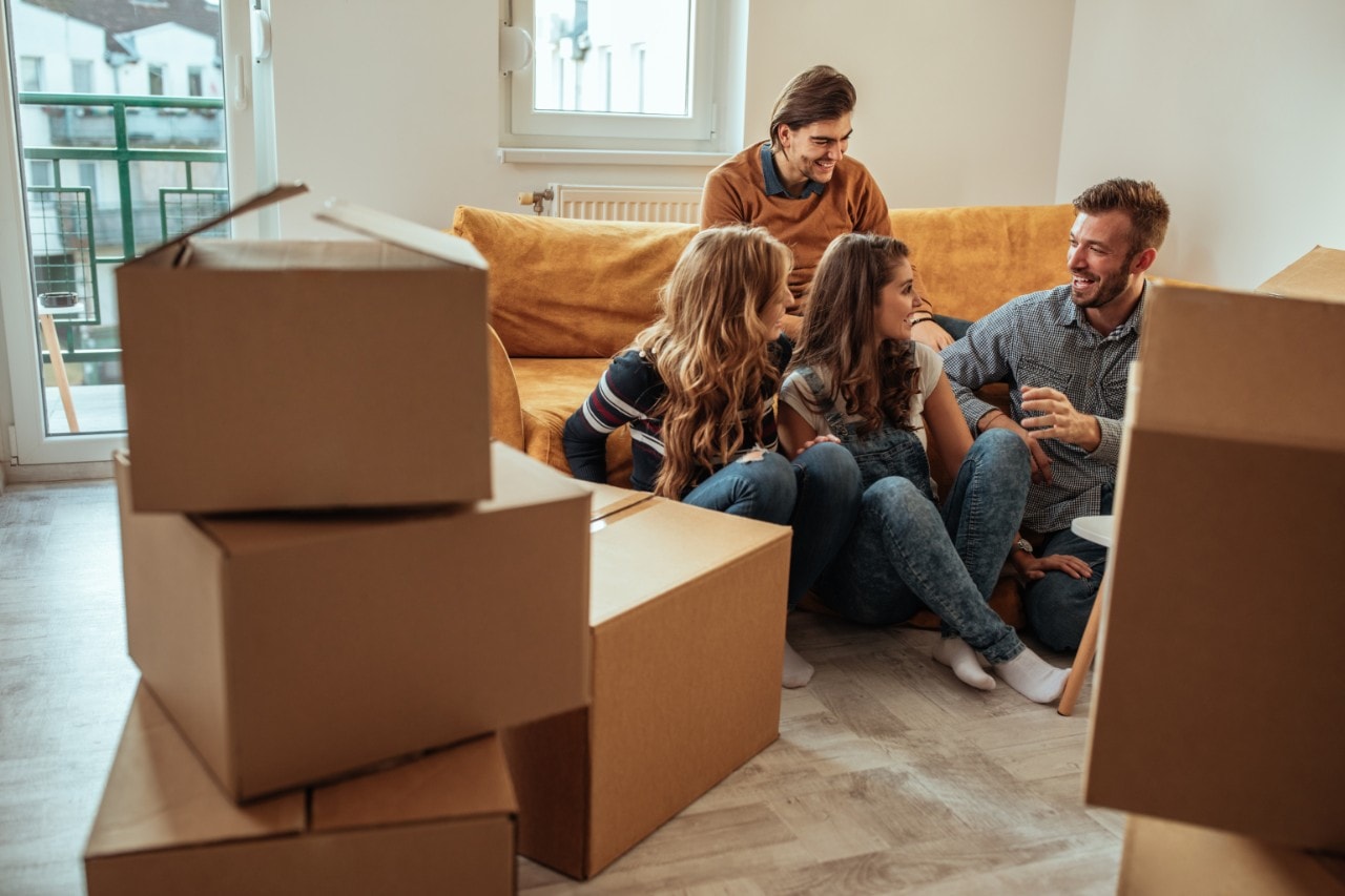 A group of people sitting around a couch, surrounded by cardboard boxes like they just moved in or are moving out.