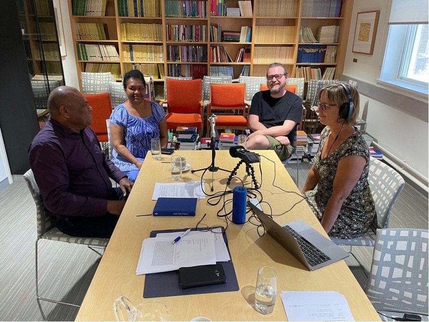 A group of four people gathered around recording equipment recording a podcast together