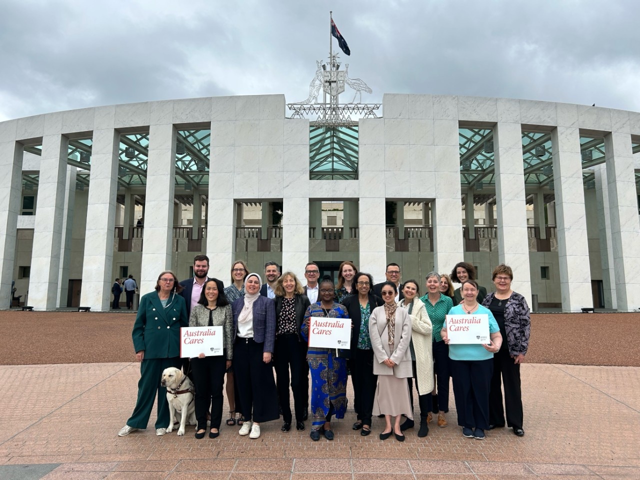 A group of people stand in front of Parliament House in Canberra holding signs that read "Australia Cares"