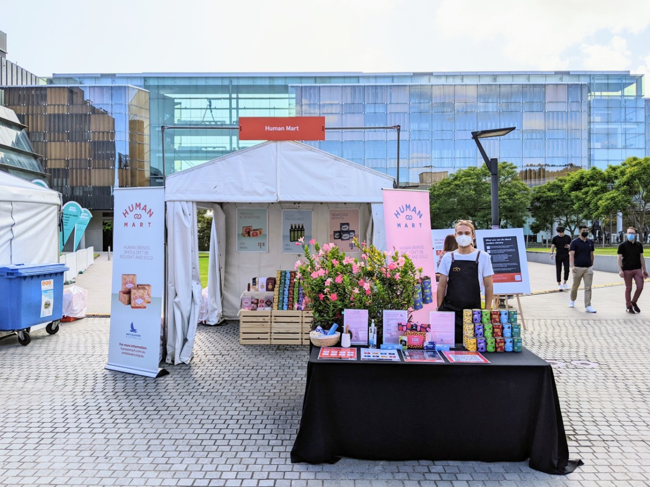 A photograph of the 'Human Mart' stall on Eastern Avenue, featuring a desk in front of the stall with empty products. Josh is standing behind the desk with wearing an apron and face mask.