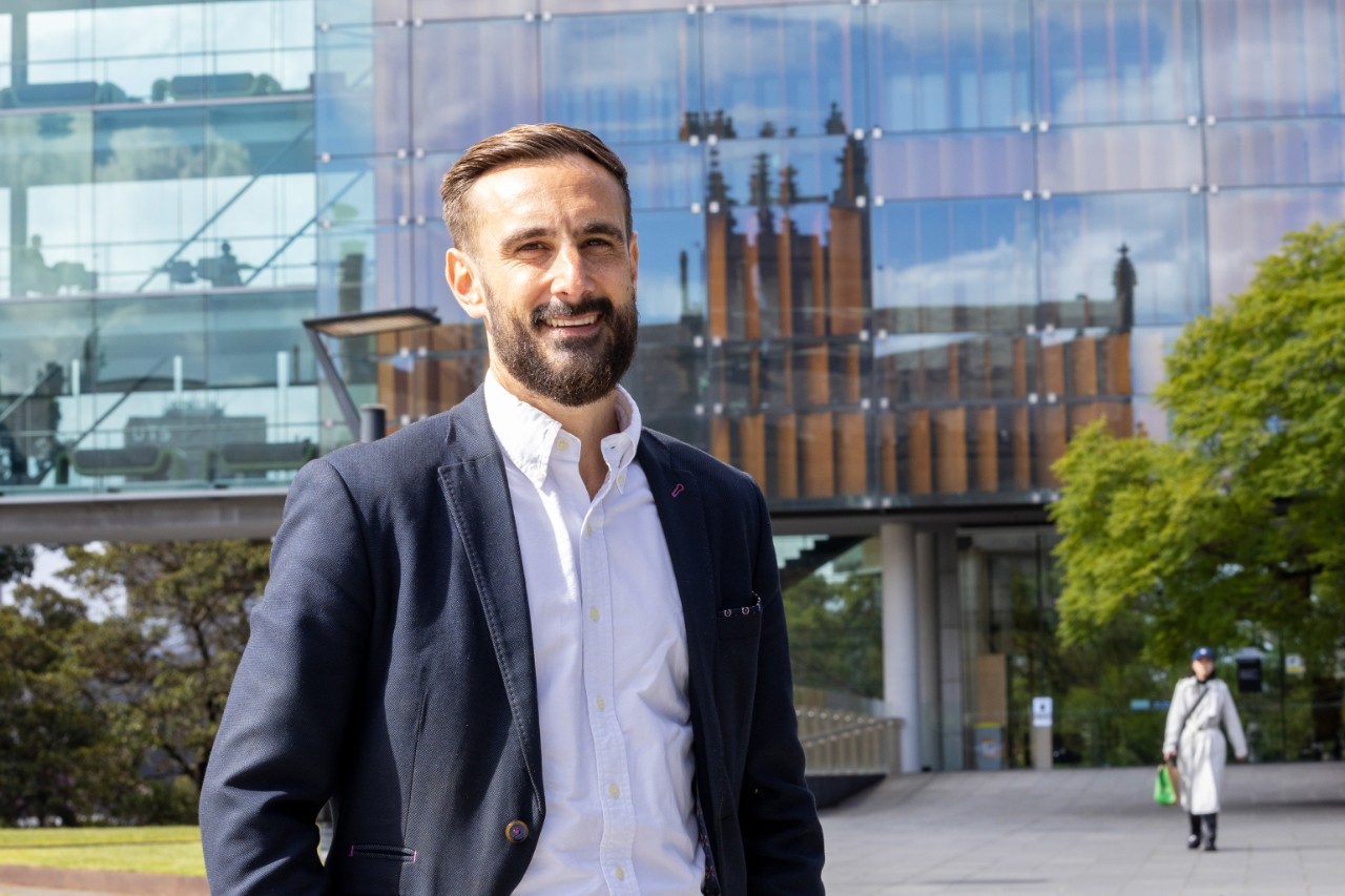 Mathew Demetriou standing in front of the Law School in a suit, smiling.