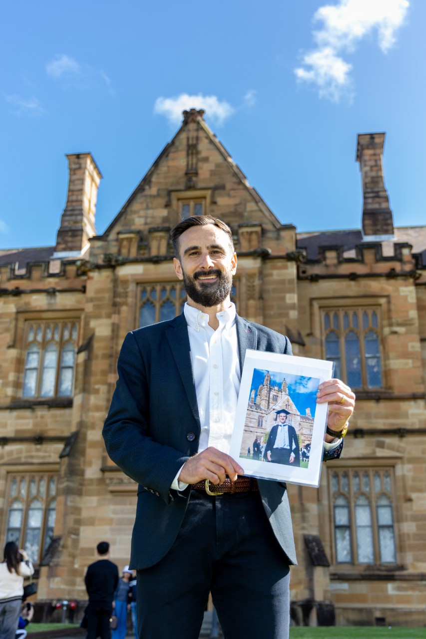 Mathew Demetriou in front of the Quad staring down at the camera holding his graduation photo, which was taken in a similar position. 