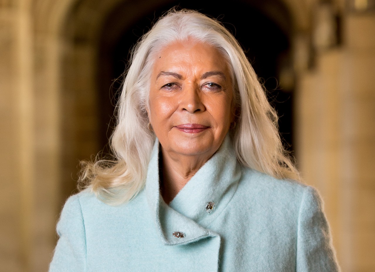Marcia Langton wearing a teal coat stands in front of a blurred sand coloured hallway