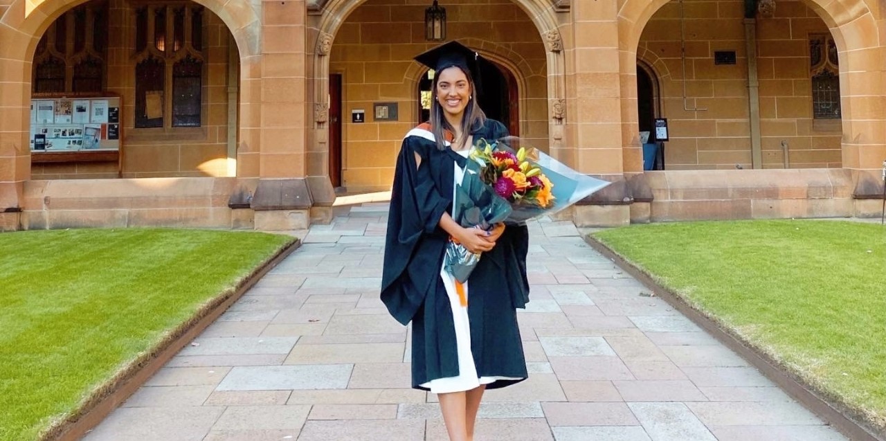 Elysee Dubois standing in front of quad holding flowers and wearing a graduation gown
