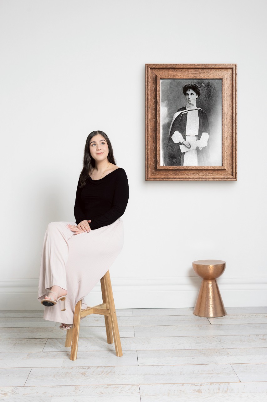 Nora sitting on a chair with a frame in the background, insluding a black and white photograph of Ada Evans