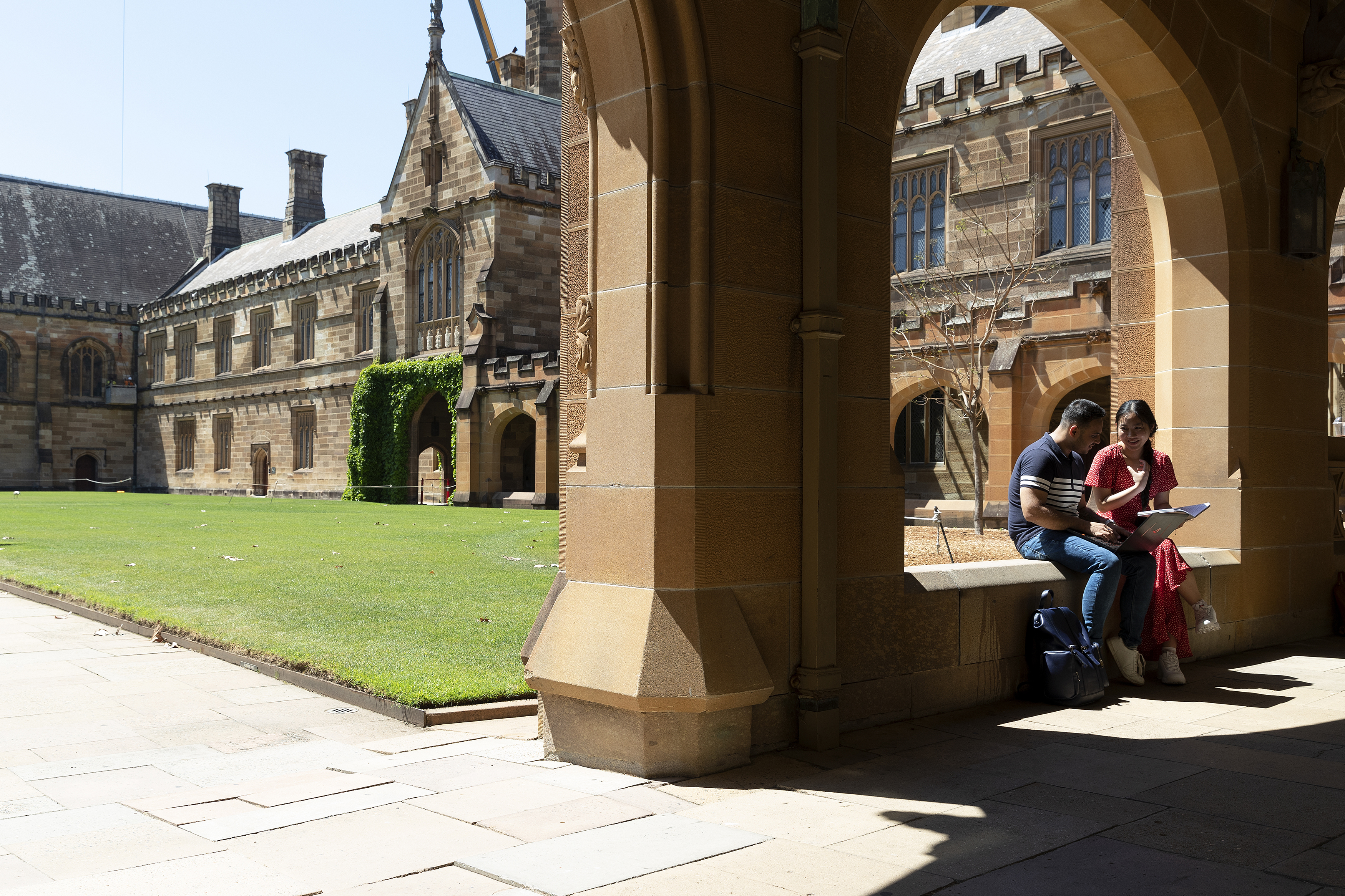 A young man and a young woman sit together under a sandstone archway in the University of Sydney Quad