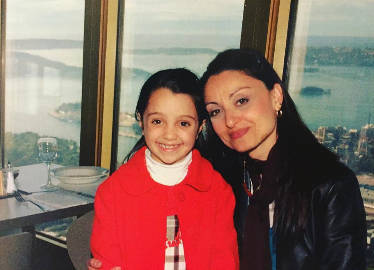 Piadora wearing a pink dress and a black graduation cloak and cap, standing side by side with her mother, smiling.