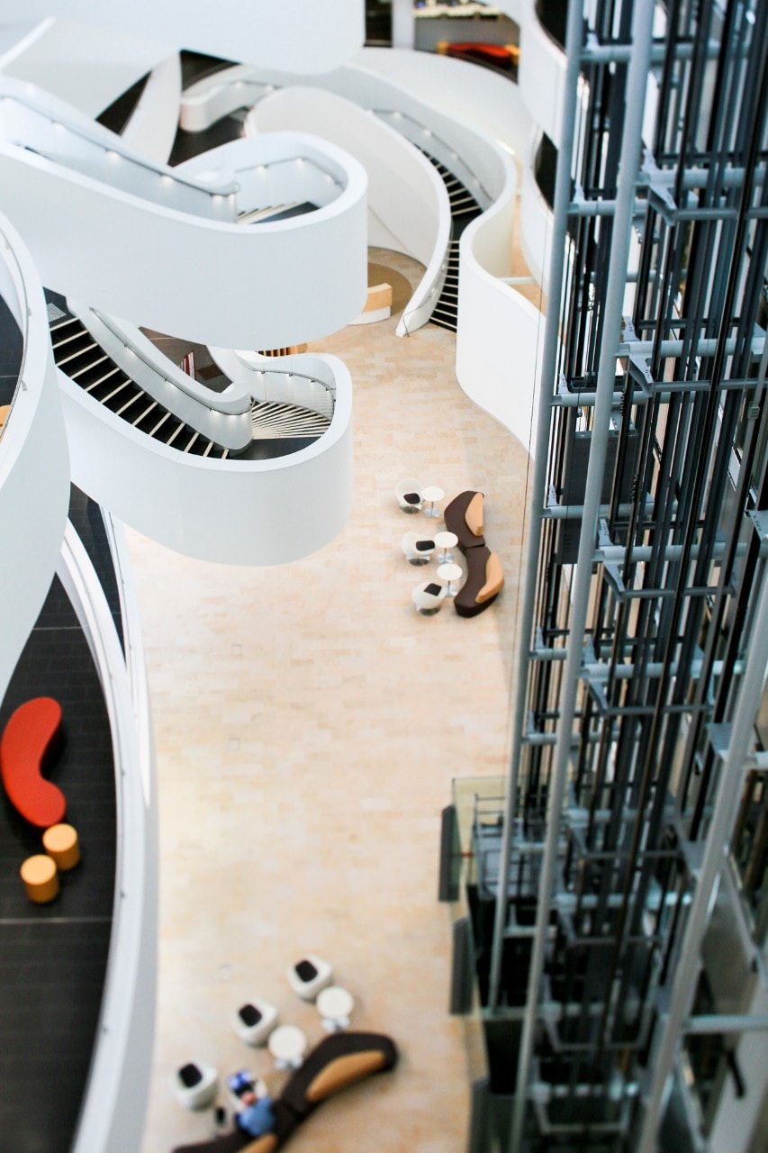 Indoors of the CPC from the top down, a white spiralling staircase on the left, a grey metallic elevator structure on the right, and a light wooden ground with people standing.