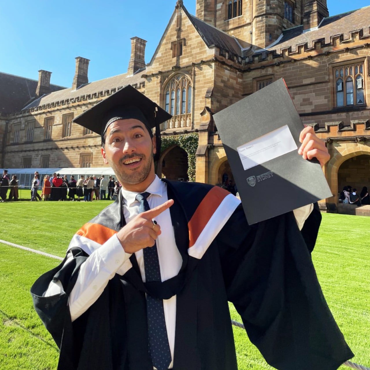 Yaser in his graduation gown holds up his degree certificate, standing on the grass in the University of Sydney's Main Quadrangle.