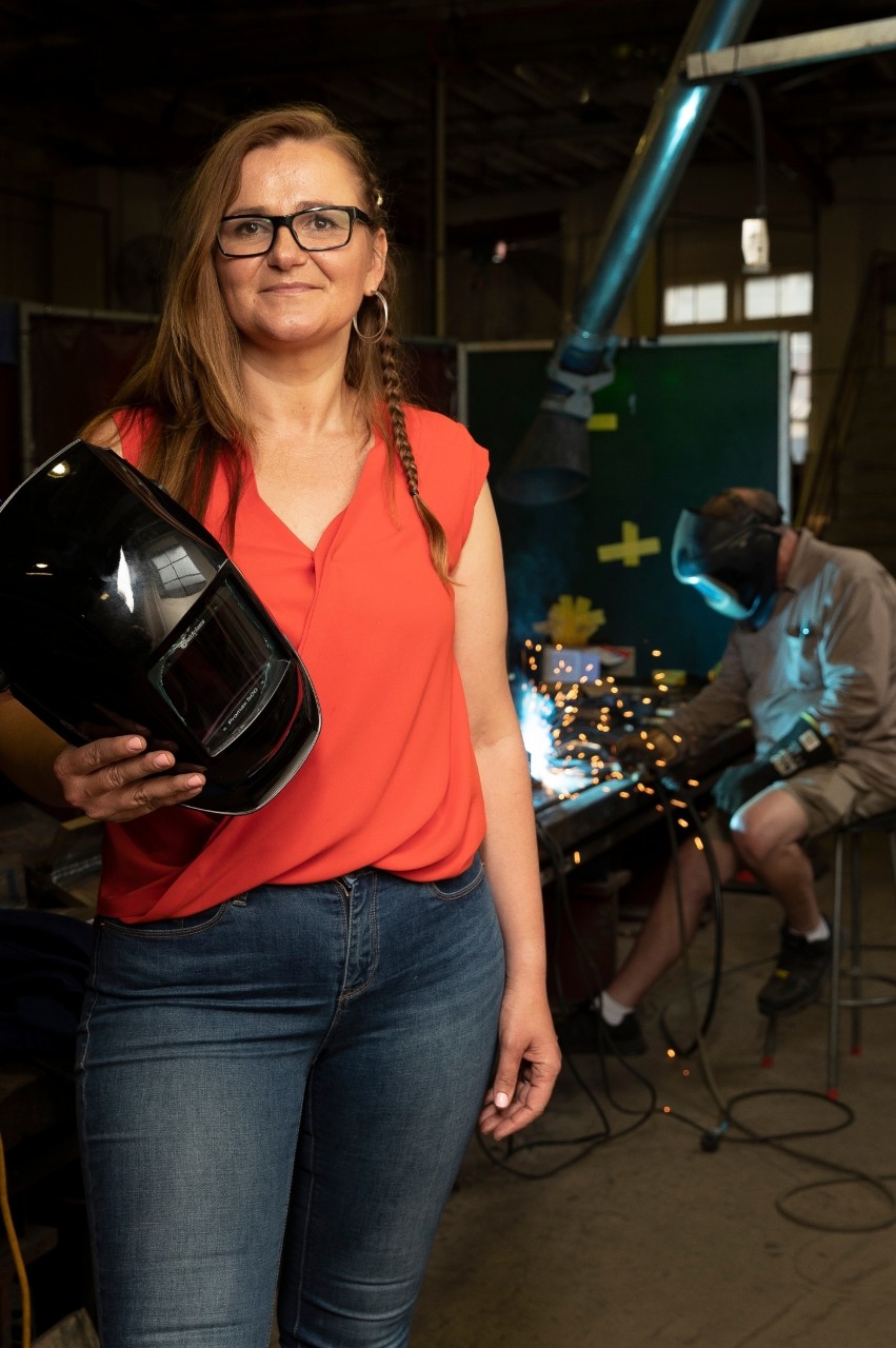 Image of Anna Ziara-Paradowska smiling for the camera with welding sparks lighting up the background behind her. 