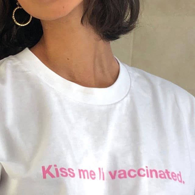 A girl (face not shown), wearing a white T-shirt that has the sentence 'Kiss me I'm vaccianted' in pink written on it