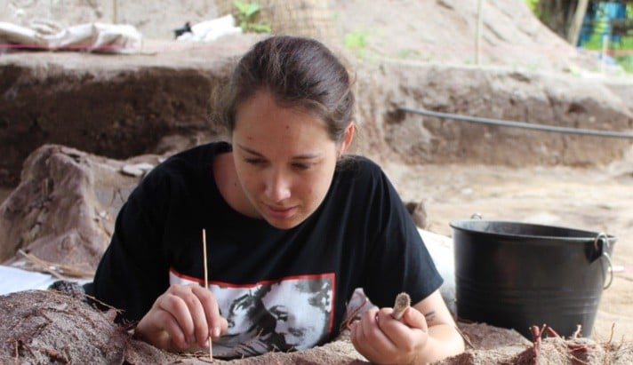 Dr Melandri Vlok in a black tshirt and hair in ponytail. She is looking down at an archaeological dig with a paintbrush in one hand
