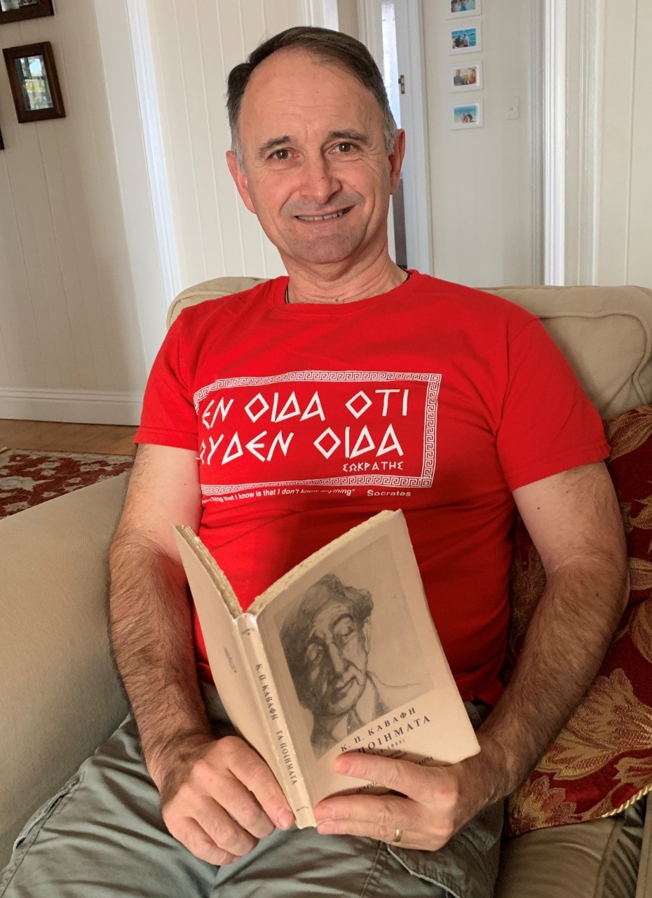 Man sitting on a sofa chair at home with a red t-shirt with Greek text and an opened Modern Greek language book in his hands resting on his laps