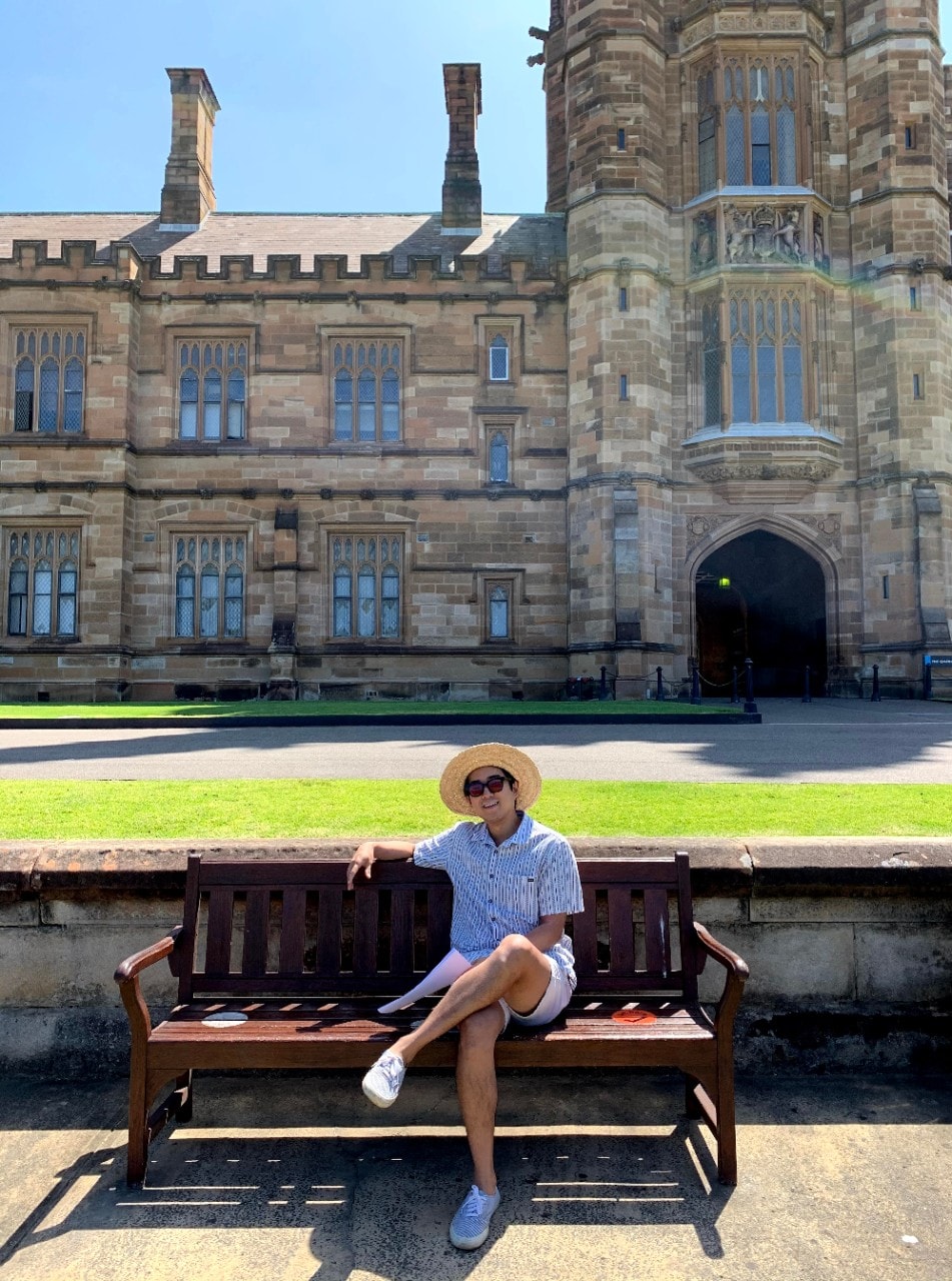 Paul Whang sitting on a wooden bench at the University of Sydney quadrangle