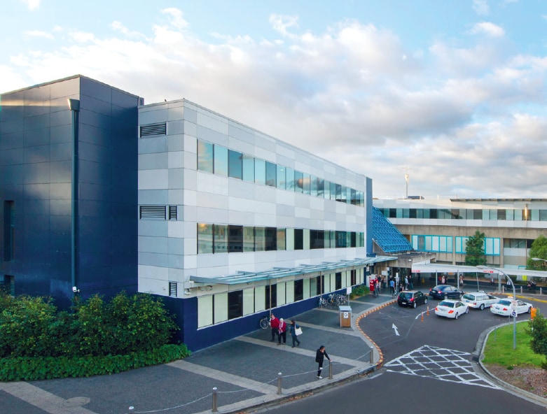 White tiled exterior view of Westmead Hospital, blue sky and clouds with busy road and pathway